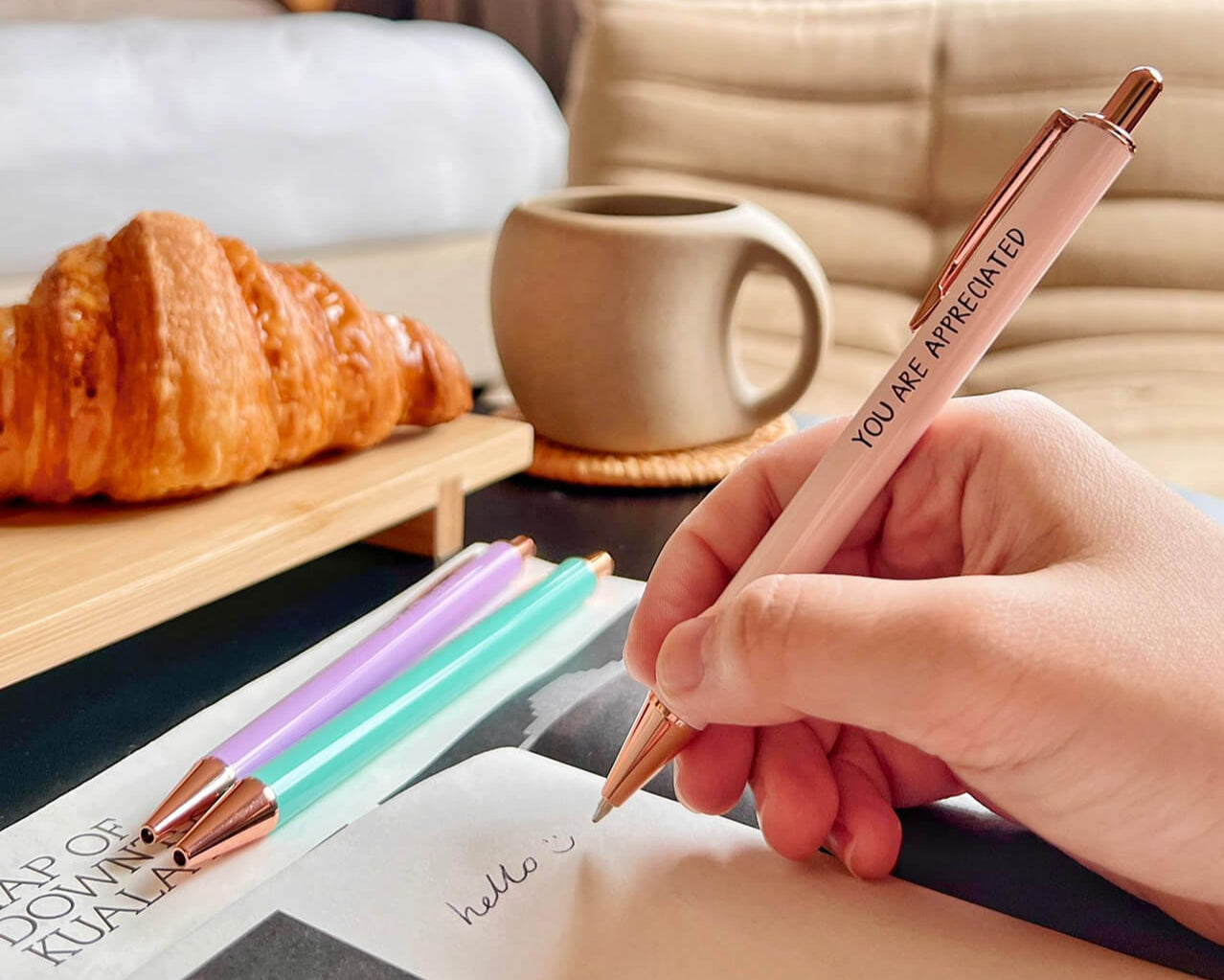 Person holding a pen with 'You are appreciated' message, next to croissants and a mug on a table.